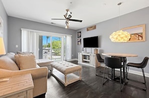 A modern living room with a sofa, ottoman, dining table, chairs, and a ceiling fan.