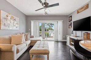 A modern living room with a sofa, ottoman, ceiling fan, TV, and a large window with a view. - NEWLY RENOVATED 2BR Golfer's Paradise Condo at World Tour (Myrtle Beach)