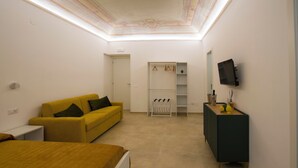 Une chambre d’hôtel avec un canapé jaune, un lit, un téléviseur fixé au mur et une petite commode avec un miroir.