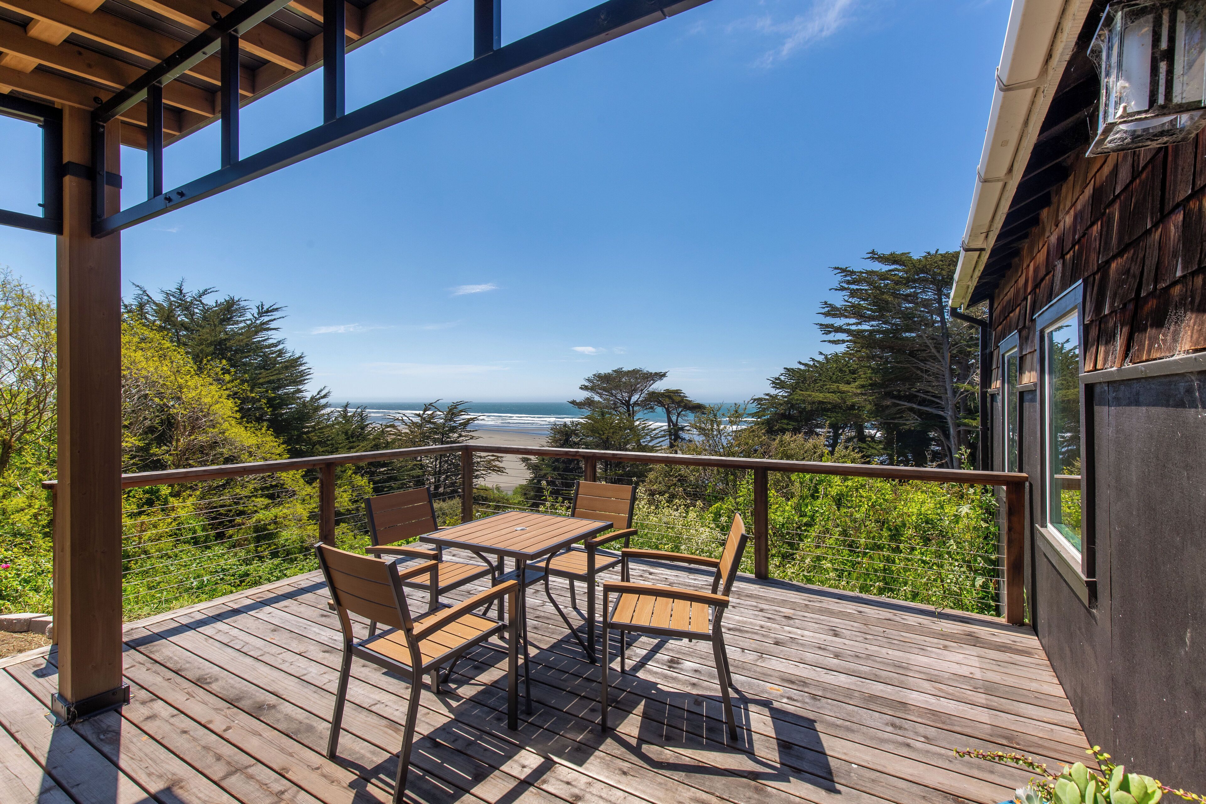 A wooden deck with a table and chairs, overlooking a beach and ocean.