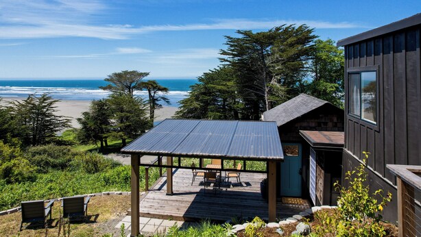 A wooden deck with a metal roof overlooking a beach, surrounded by trees and a small building with a blue door.