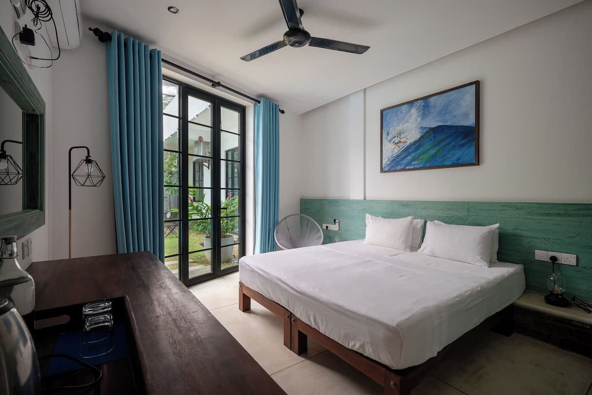 A hotel room with a bed, a desk, a ceiling fan, a painting, and a view of the outdoors.