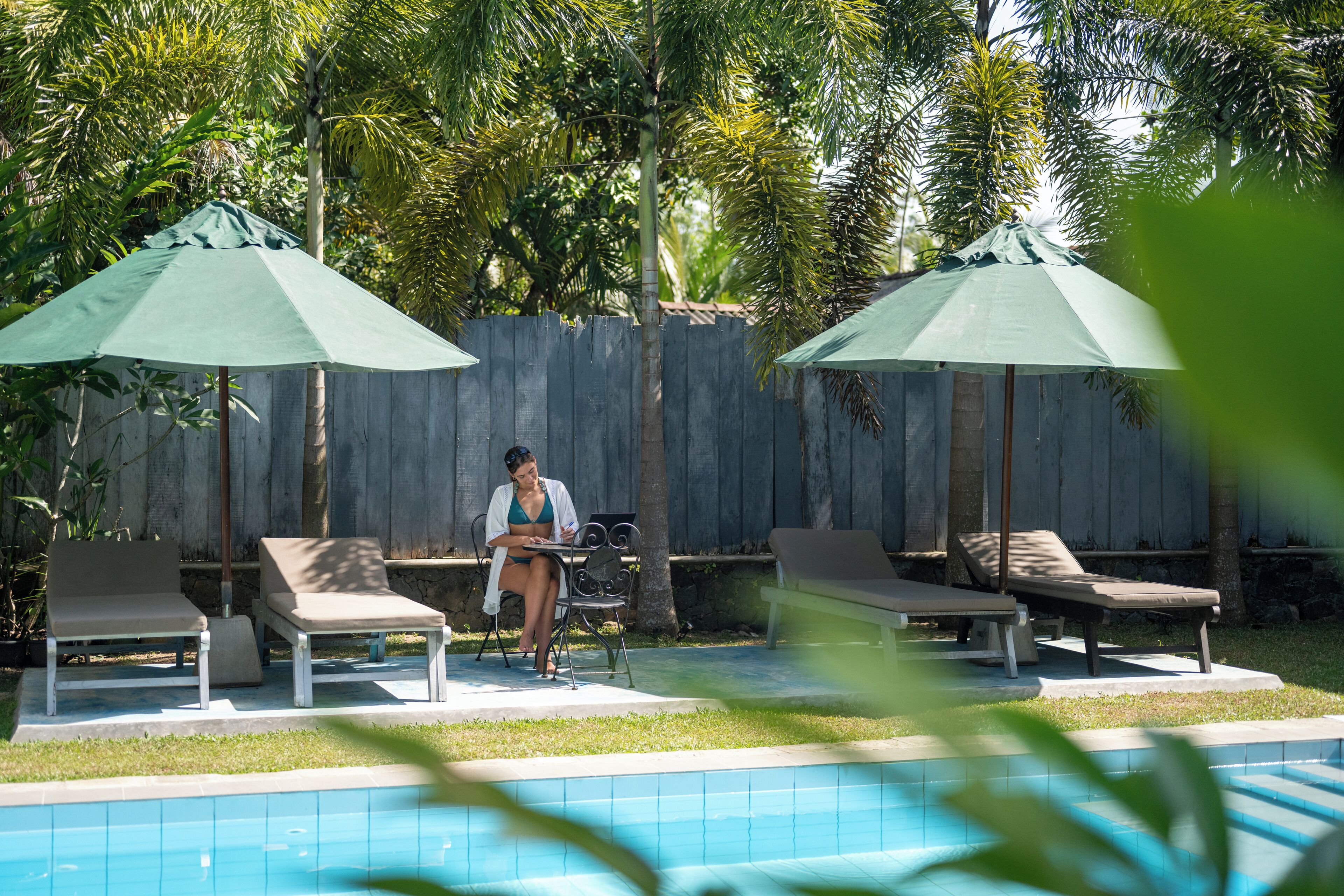 Un área junto a la piscina con sillones de descanso y sombrillas, una persona sentada a una mesa y una cerca de madera al fondo.