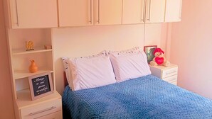 A bedroom with a bed, white cabinets, a small shelf with decorative items, and a bedside table with a teddy bear.