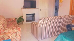 A living room with a sofa, a striped armchair, a television above a fireplace, and a dining table with a green tablecloth.