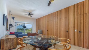 Un salon moderne avec une table à manger au plateau en verre, entourée de chaises en osier, un mur d’accent en bois et un ventilateur de plafond.