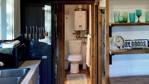 Cocina compacta con lavabo, estufa y armarios, que da paso a un baño con tanque de agua a la vista.