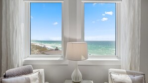 A room with two windows overlooking the ocean, a lamp on a table, and two chairs with cushions.
