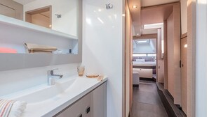 Un pasillo estrecho y bien iluminado con un lavamanos y un espejo blancos, que da a un dormitorio con cama y ventana.