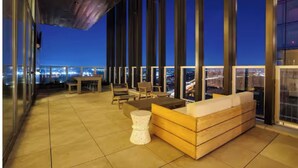 A modern outdoor lounge area with a high-rise city view, featuring a wooden bench, a small round table, and a seating arrangement with chairs. - Luxury Rainey St Condo with rooftop pool, Lakeview & Gym (Austin)