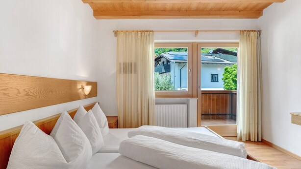 Ein Schlafzimmer mit hölzernem Kopfteil, einem Bett mit weißer Bettwäsche, einem Fenster mit Vorhängen und Blick auf ein Gebäude und Grünfläche.