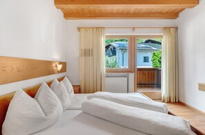 A bedroom with a wooden headboard, a bed with white bedding, a window with curtains, and a view of a building and greenery outside. - Apartment 'Hofanger Meran' with Mountain View, Balcony and Wi-Fi (Algund)
