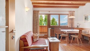 Ein gemütliches Zimmer mit Holzbalkendecke, einer Couch mit Kissen, einem Tisch mit Stühlen und einem Fenster mit Vorhängen.
