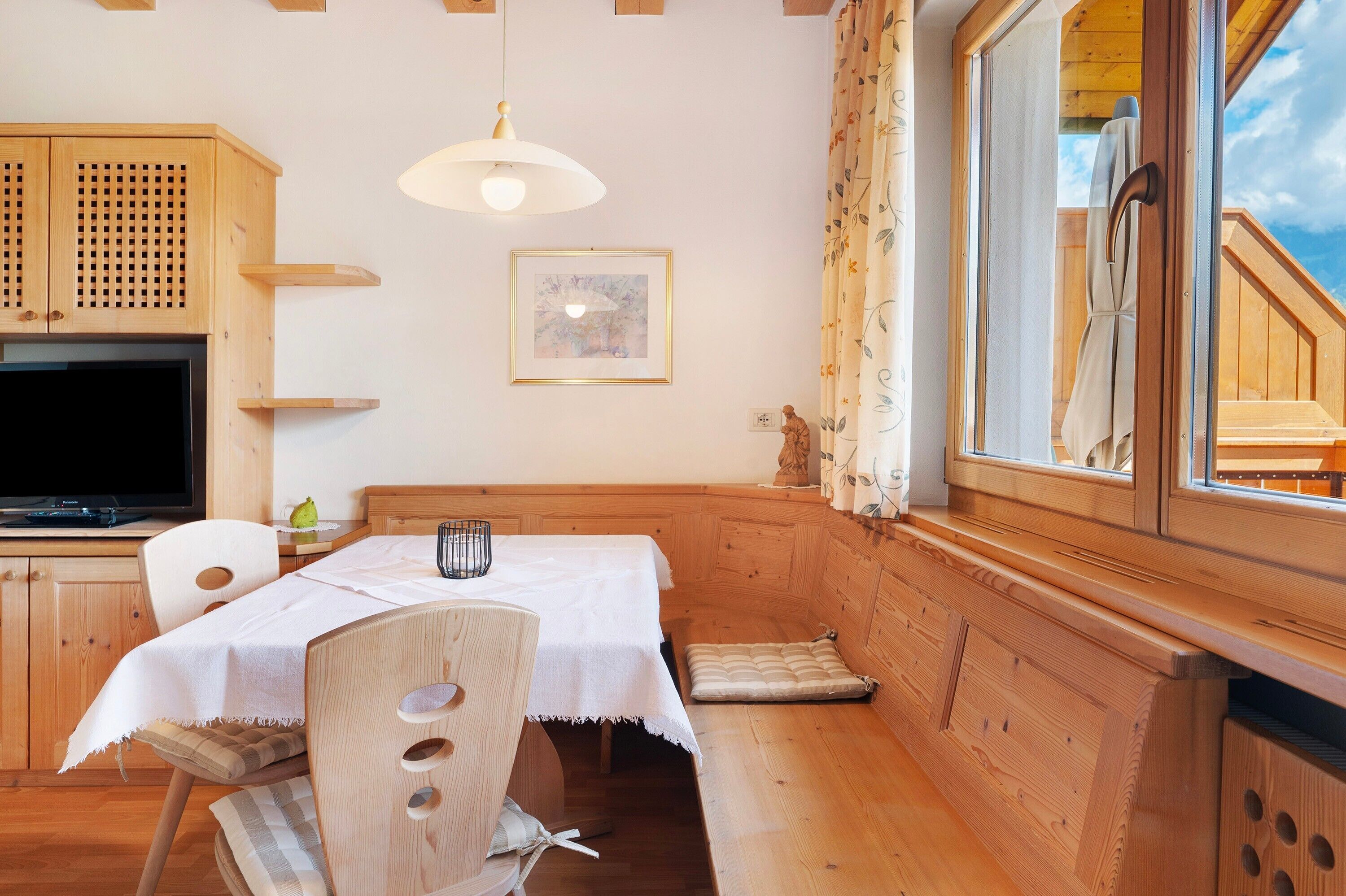 A wooden cabin interior with a dining table, chairs, a television, a framed picture, and a window with curtains.