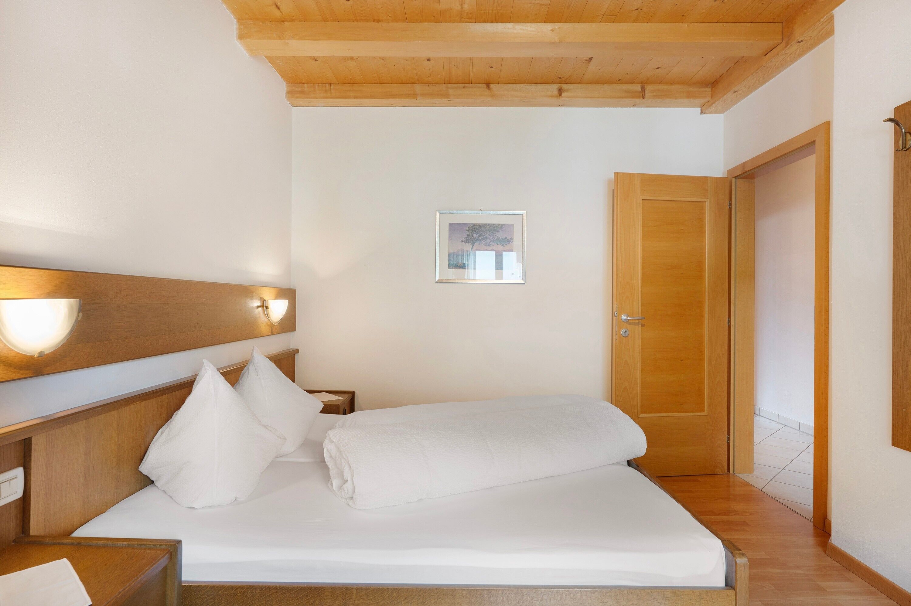 A neatly made bed with white linens, a wooden headboard, a bedside table with a lamp, and a framed picture on the wall.
