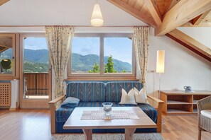 Une pièce avec un plafond en bois, un canapé agrémenté de coussins à motifs, une table sur laquelle se trouve un vase en verre, et une vue sur les montagnes par de grandes fenêtres.
