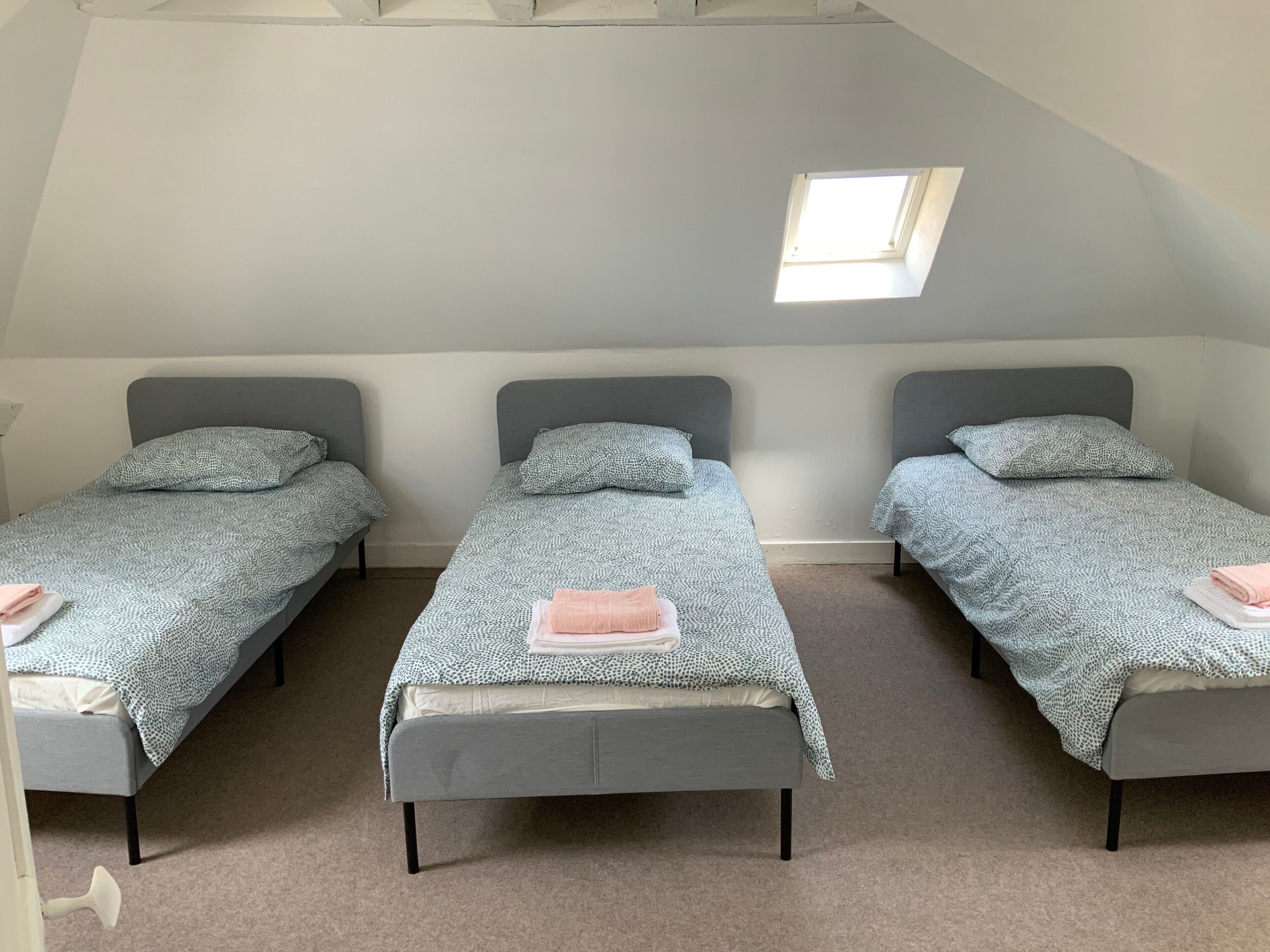 Una stanza con tre letti singoli, ognuno con un poggiatesta grigio e una coperta a motivi decorativi. Soffitta con finestra a lucernario sopra il letto centrale e soffitto a falde.