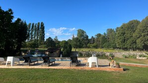 Un espace piscine avec des chaises longues, un petit bâtiment et un paysage verdoyant parsemé d’arbres.