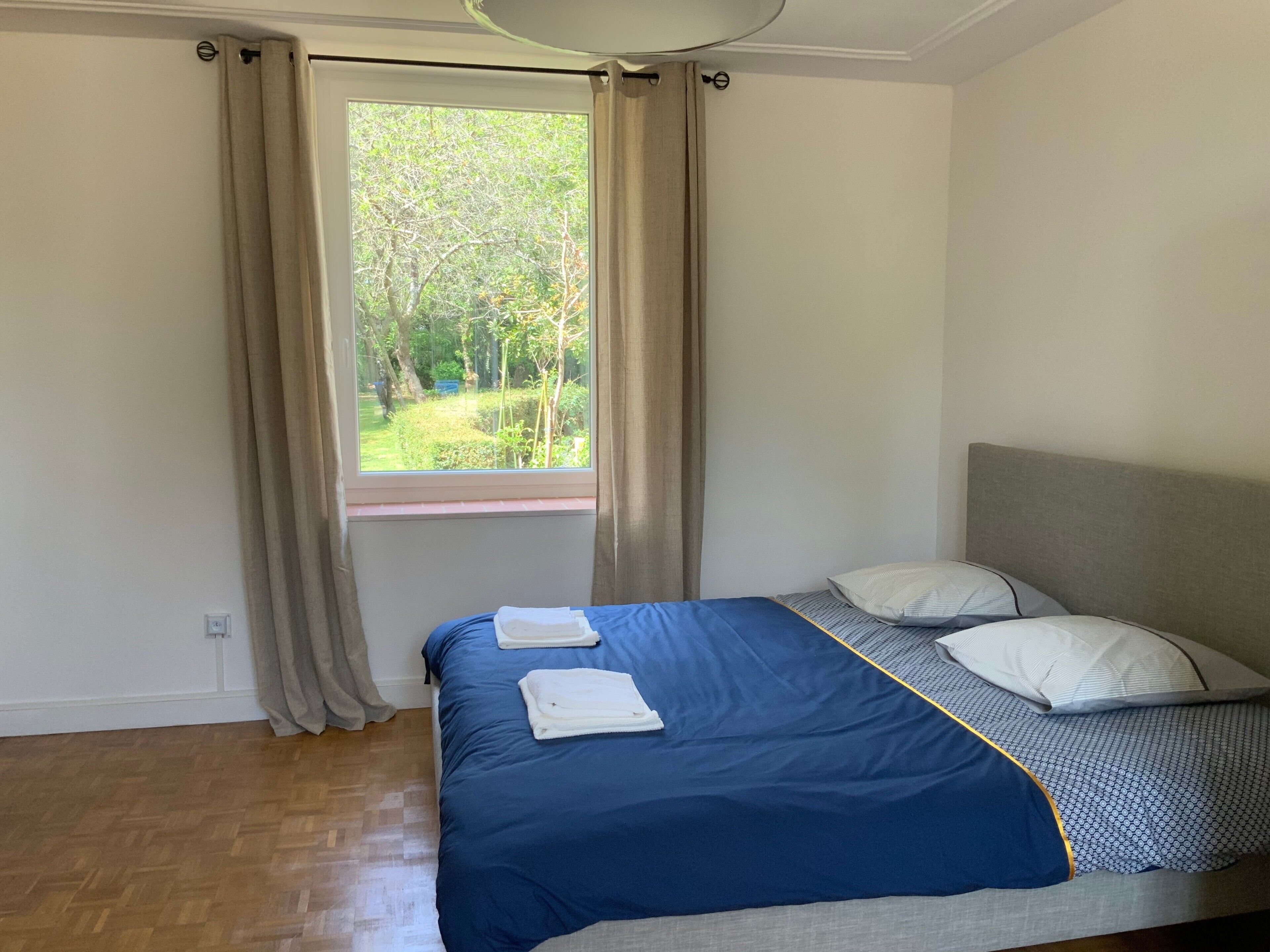 Een slaapkamer met een bed, gordijnen, een raam met uitzicht op groen en een houten vloer.