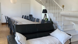 Un salon moderne avec un canapé noir, des coussins blancs, une table à manger avec des chaises, un escalier et un luminaire suspendu.