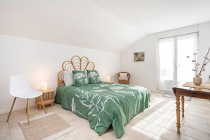 A bedroom with a green floral bedspread, a white chair, a wooden bedside table, and a large window.