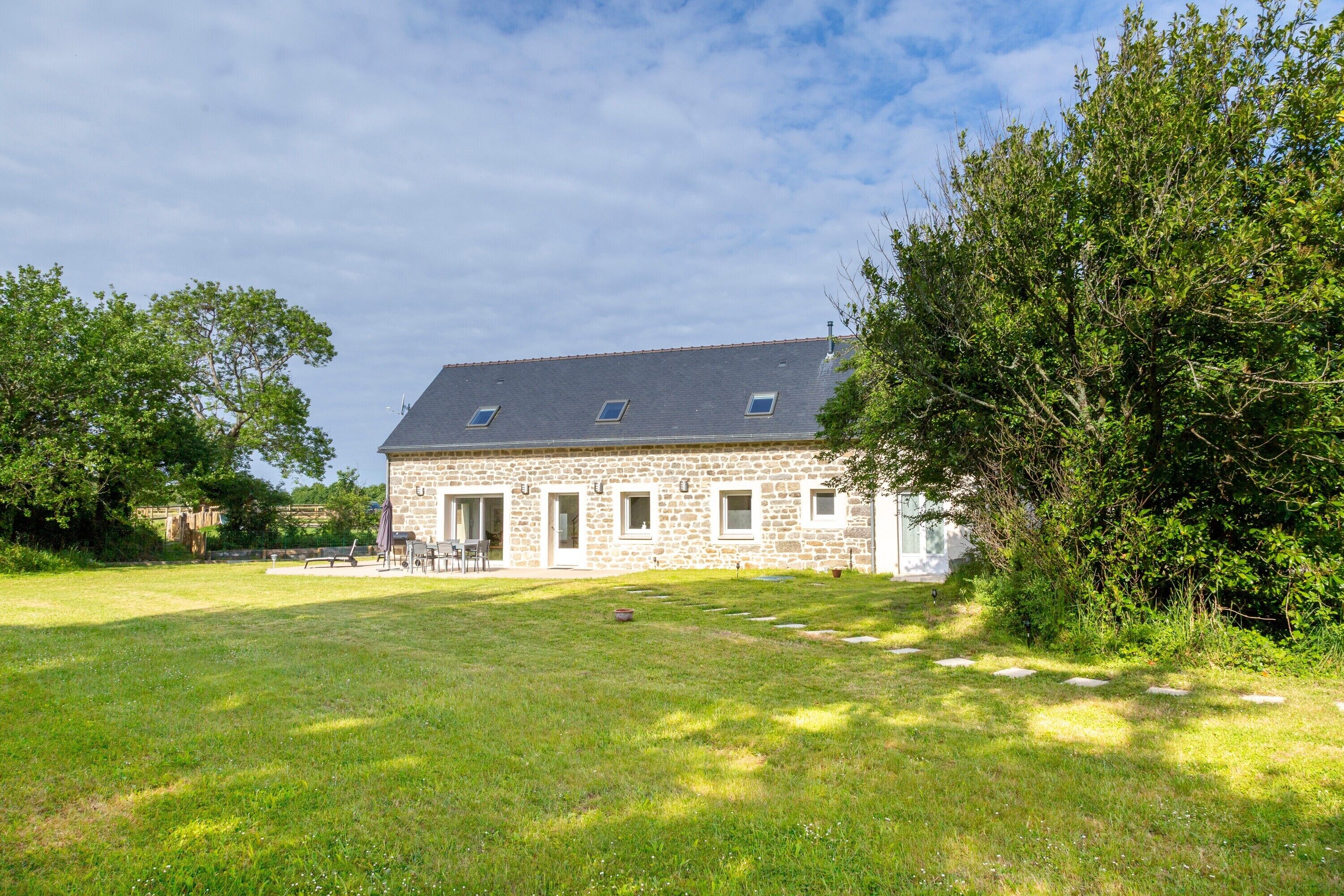 Een gebouw van steen met een schuin dak, een gazon en een grote boom aan de rechterkant.