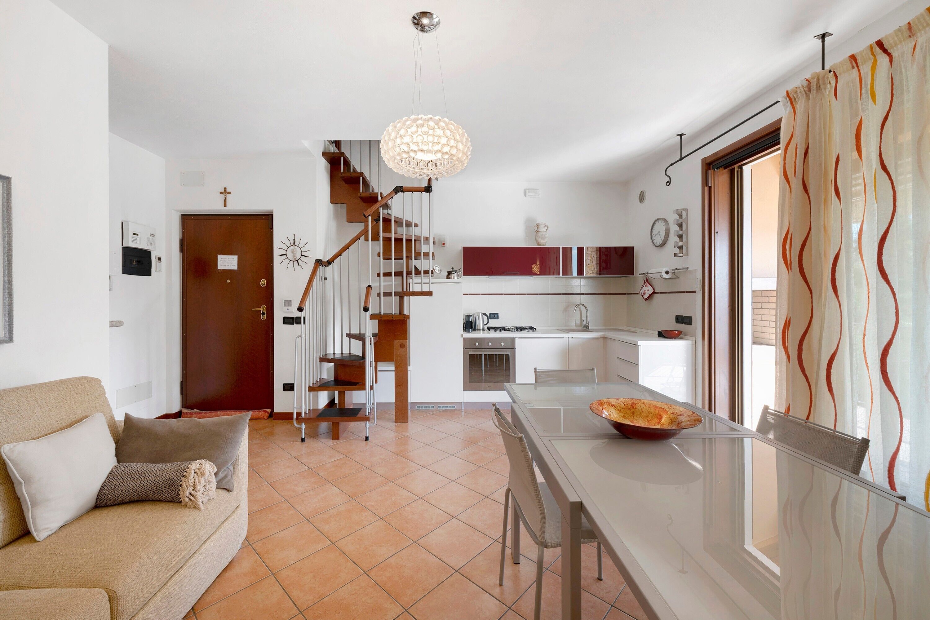 A modern kitchen with a central island, a dining area with a glass table, a staircase leading to a loft, and a living room with a sofa.