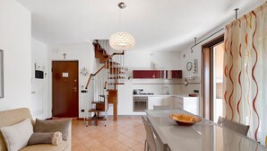 A modern kitchen with a central island, a dining area with a glass table, a staircase leading to a loft, and a living room with a sofa.