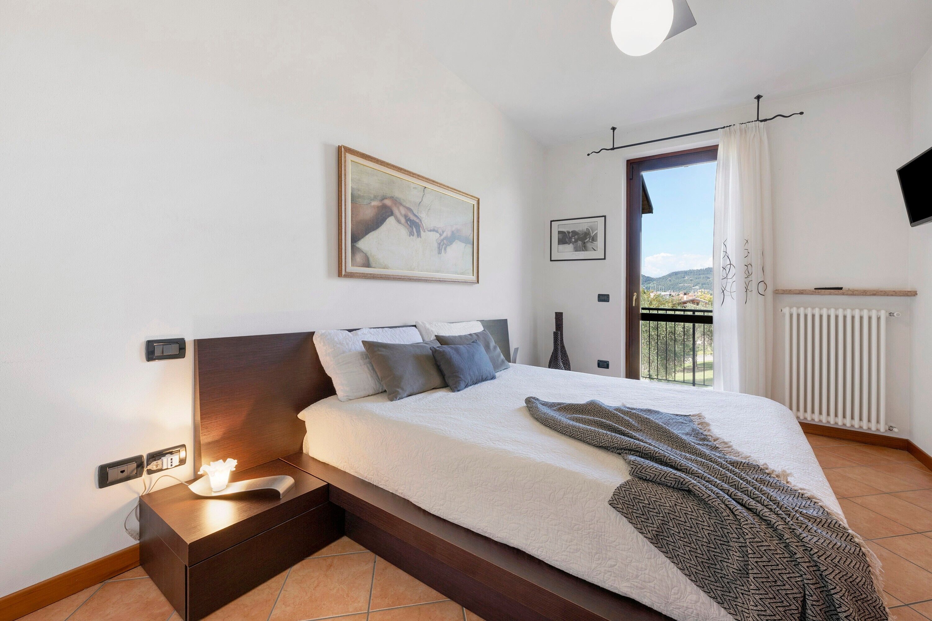 A bedroom with a bed, bedside table, wall art, and a view through a window.