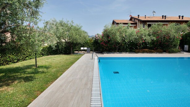 A swimming pool with a tiled deck, surrounded by greenery and a building with a tiled roof.