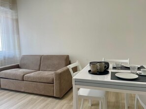 Una sala moderna con un sofá color beige, una mesa de comedor blanca con dos sillas y una ventana con cortinas translúcidas.