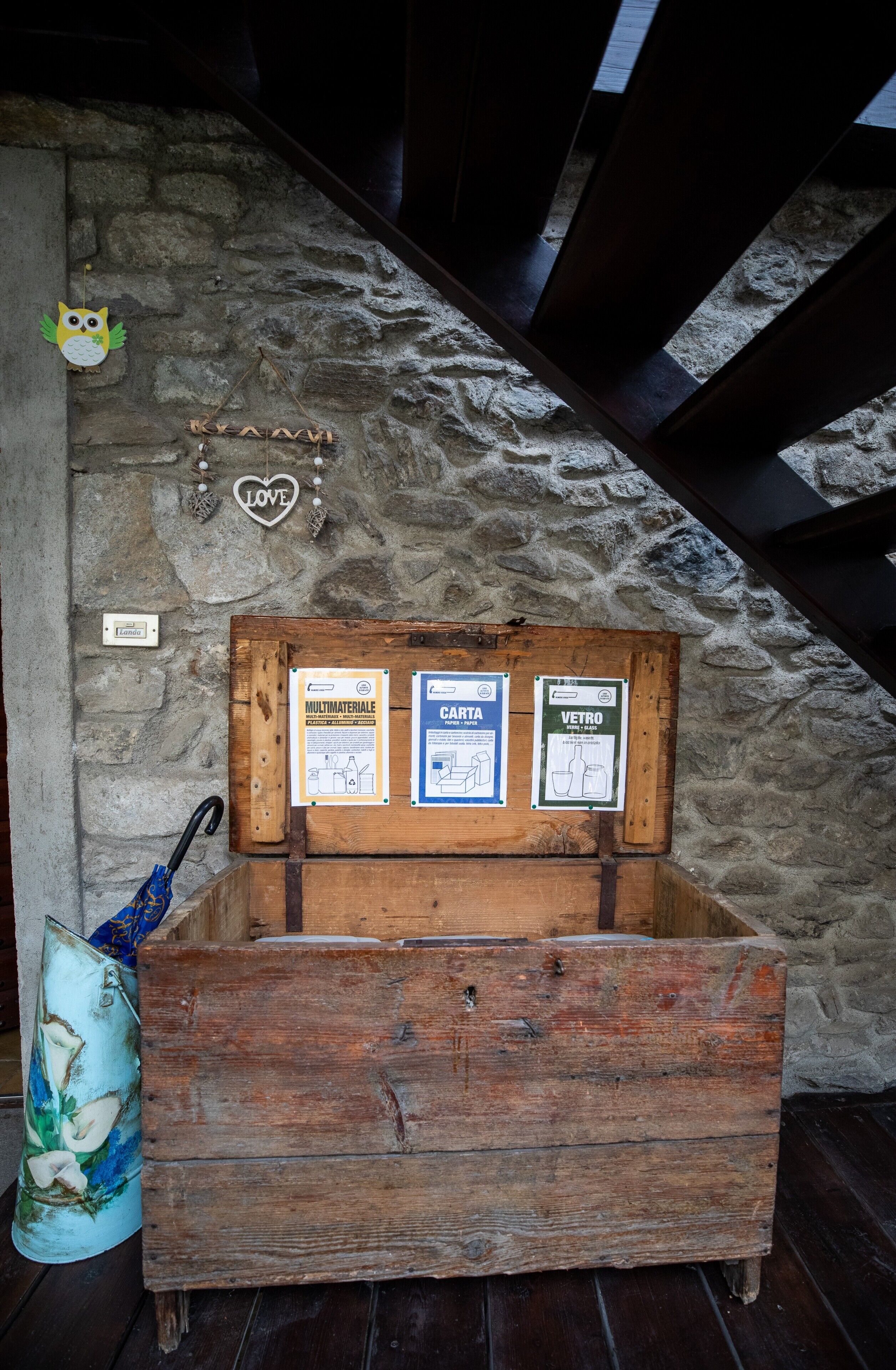 A rustic wooden chest with three informational signs against a stone wall, a small owl decoration, and a heart-shaped sign.