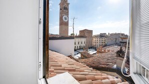 Vista da janela sobre uma paisagem urbana com uma torre do relógio imponente, telhados de terracota e uma mistura de edifícios históricos e modernos.