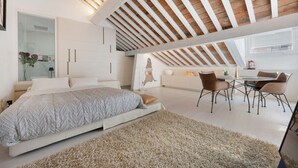 Quarto com cama, mesa de jantar com cadeiras, sofá branco e teto com vigas de madeira.