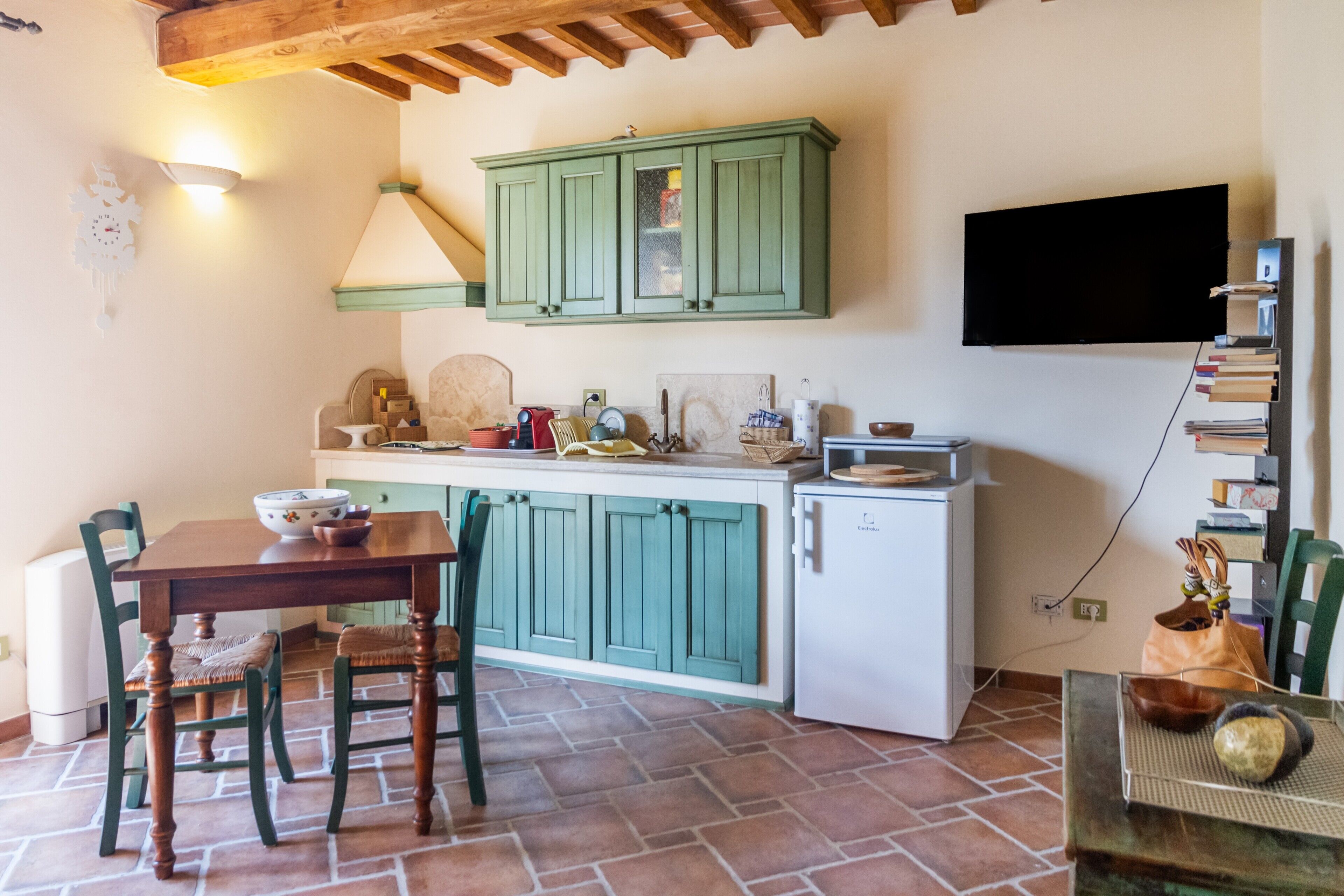 Una cucina con un tavolo di legno, due sedie, un armadietto verde, un frigorifero bianco e una TV a parete.