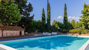 Une piscine aux eaux cristallines, bordée de chaises longues blanches, entourée d’arbres et d’un bâtiment en pierre.