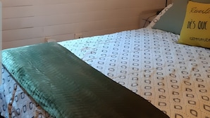 Une chambre avec un plafond en pente, un tableau encadré au mur, un lit avec une literie à motifs, un plaid vert et un coussin orné d’un texte.