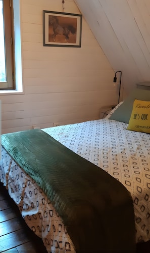 Un dormitorio con techo inclinado, un cuadro enmarcado en la pared, cama con ropa de cama estampada, una manta verde y un cojín con texto.