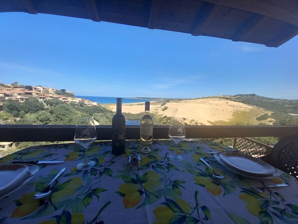 Une table dressée pour deux personnes, avec des verres à vin, une bouteille et des couverts, donnant sur un paysage côtier avec des bâtiments et des collines.