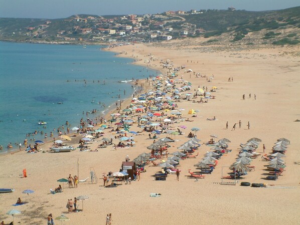 Une plage bordée de rangées de parasols et de chaises longues, un ciel dégagé et des bâtiments en arrière-plan.