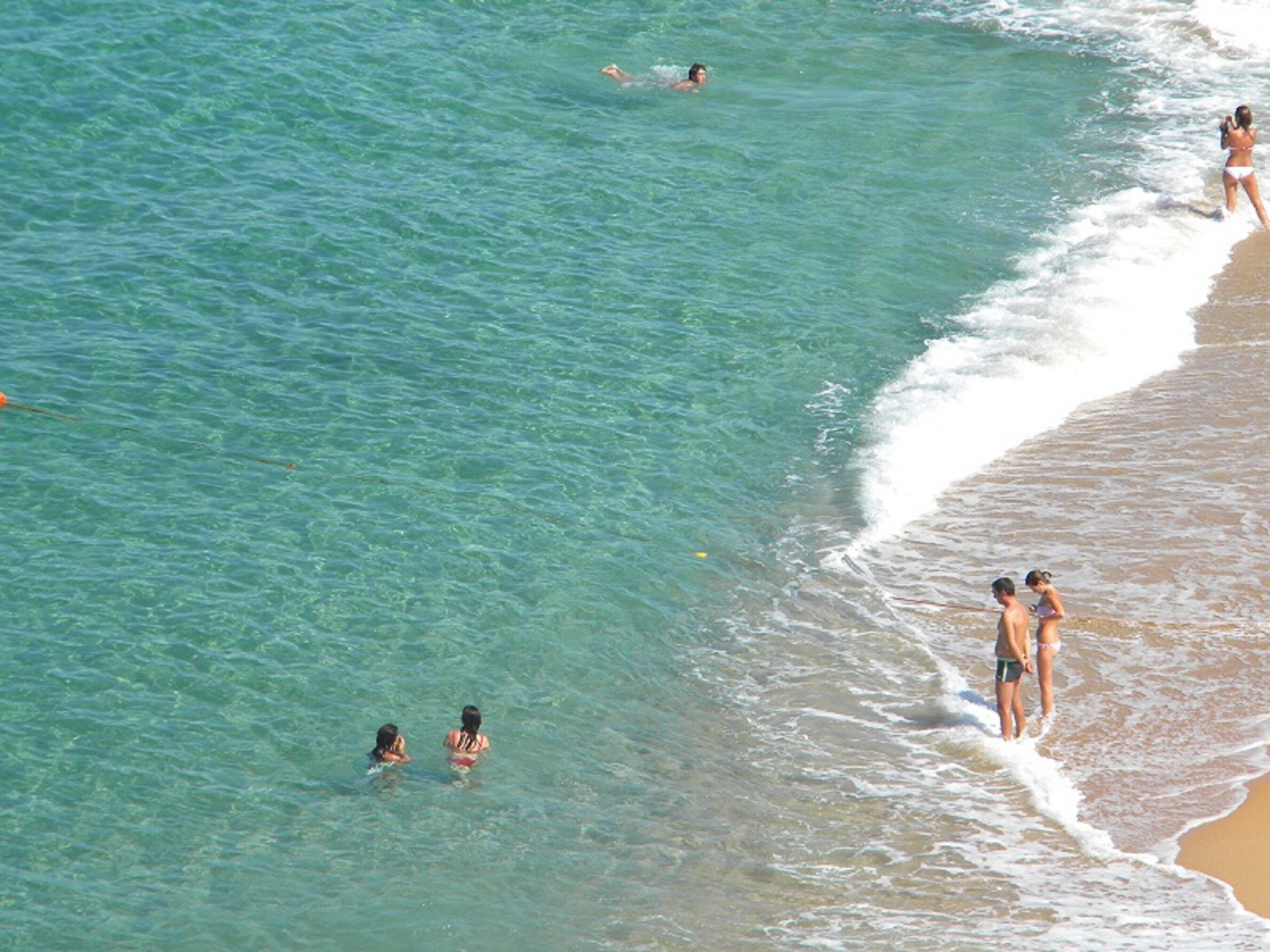 Eine Strandansicht mit klarem Wasser, Menschen schwimmen und stehen am Ufer.