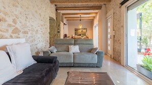 Quarto com parede de pedra, sofá, poltrona preta, porta de vidro que dá para o exterior e teto com vigas aparentes.