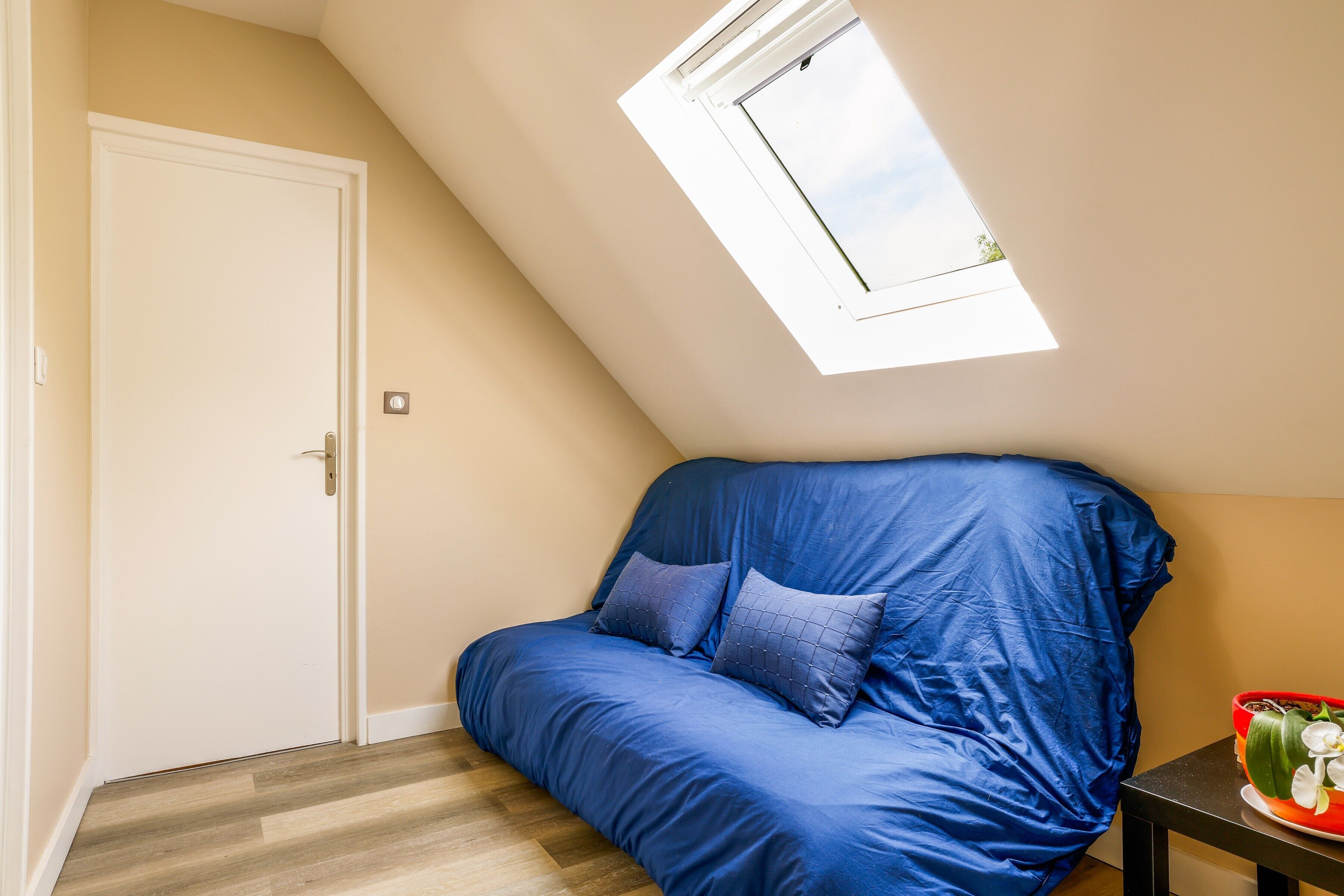Una habitación con techo inclinado, una ventana, una puerta, un sofá azul y una pequeña mesa con un cuenco rojo.