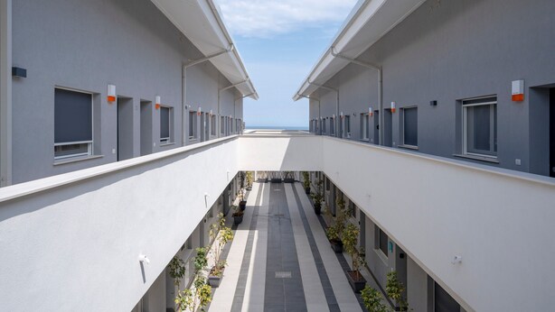 Un bâtiment moderne doté d’un couloir central, de part et d’autre duquel se trouvent des pièces, des plantes en pot, et sous lequel s’étend un ciel dégagé.