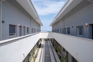 A modern building with a central corridor flanked by rooms, potted plants, and a clear sky.