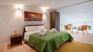 Una camera d'albergo con un letto, un comodino, una coperta verde, un quadro e un balcone con sedie in vimini.