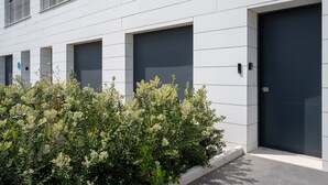 Um edifício moderno com uma fileira de portas escuras, paredes brancas e uma pequena área ajardinada com arbustos verdes.