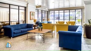 Un hall d’entrée spacieux, avec un canapé bleu, des fauteuils jaunes, une table basse et de grandes fenêtres donnant sur un paysage agréable.