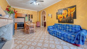 A living room with a blue sofa, a dining table with chairs, a kitchen area with cabinets, and a ceiling fan.
