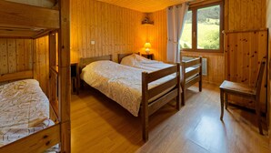 Un dormitorio tipo cabaña de madera con literas, una cama individual, una mesita de noche, una lámpara, una silla y una ventana con vistas a la vegetación.